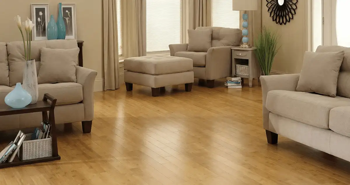A living room with professionally installed bamboo flooring, beige sofas, a matching ottoman, a side table with a blue lamp, vases, framed pictures, and large windows with blinds and curtains in Columbia, SC
